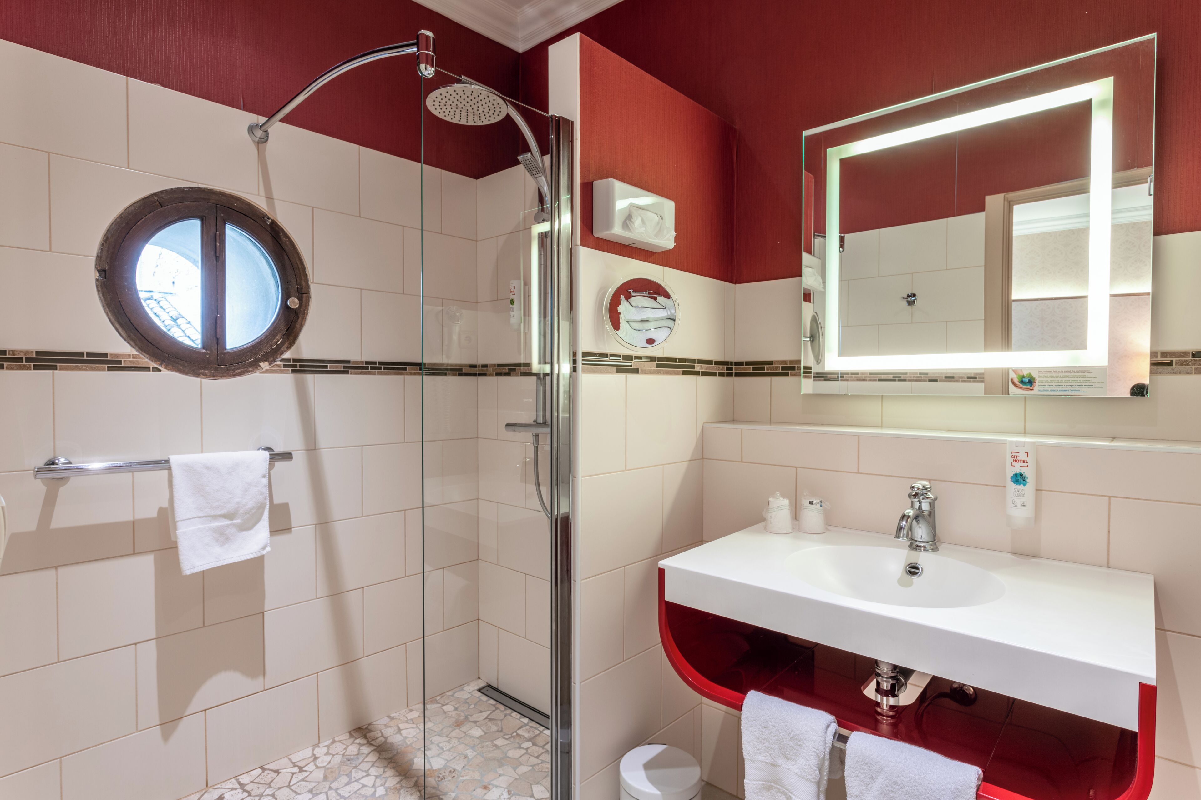 A bathroom with a glass-enclosed shower, a round mirror, a white sink, and a red accent wall.