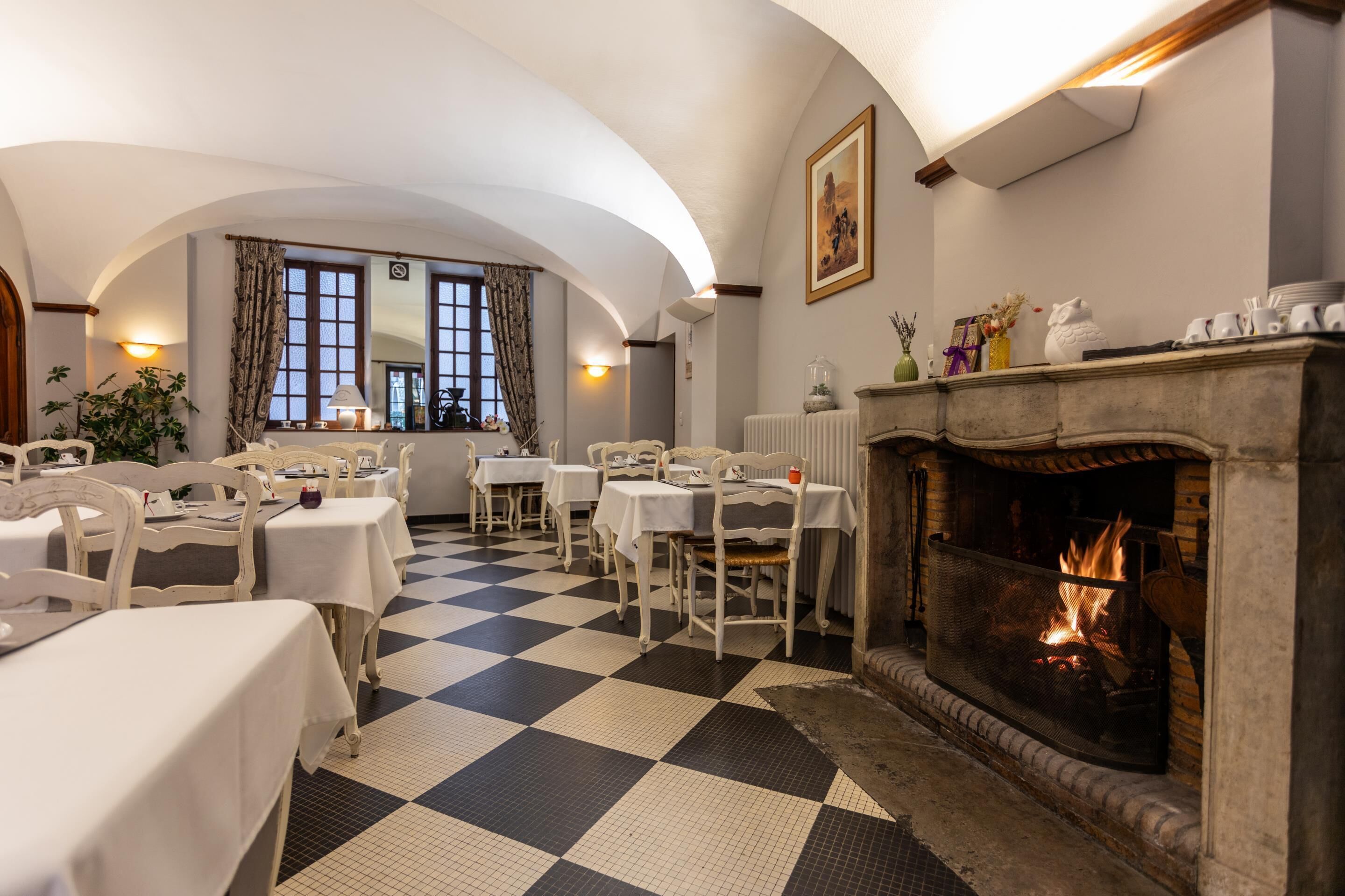 A dining area with checkered flooring, white tables, and chairs, a fireplace with a fire, and a window with curtains.