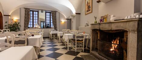 A dining area with checkered flooring, white tables, and chairs, a fireplace with a fire, and a window with curtains.