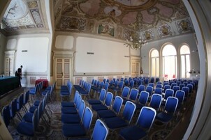 Un salón de conferencias con filas de sillas azules, un escenario con una mesa verde y detalles decorativos en el techo.