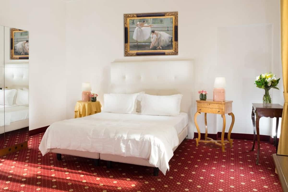 A hotel room with a large bed, a mirror, a painting, a bedside table with a lamp, and a vase with flowers.