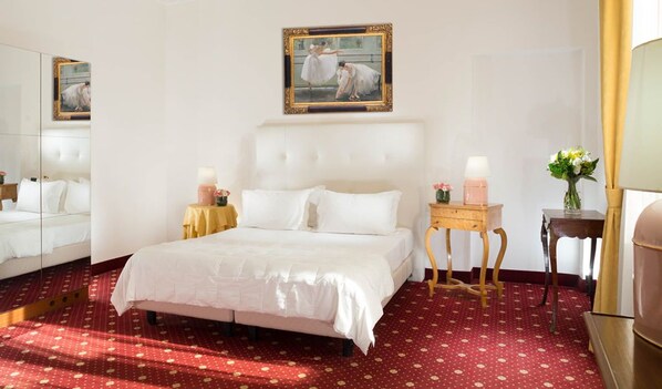 A hotel room with a large bed, a mirror, a painting, a bedside table with a lamp, and a vase with flowers.