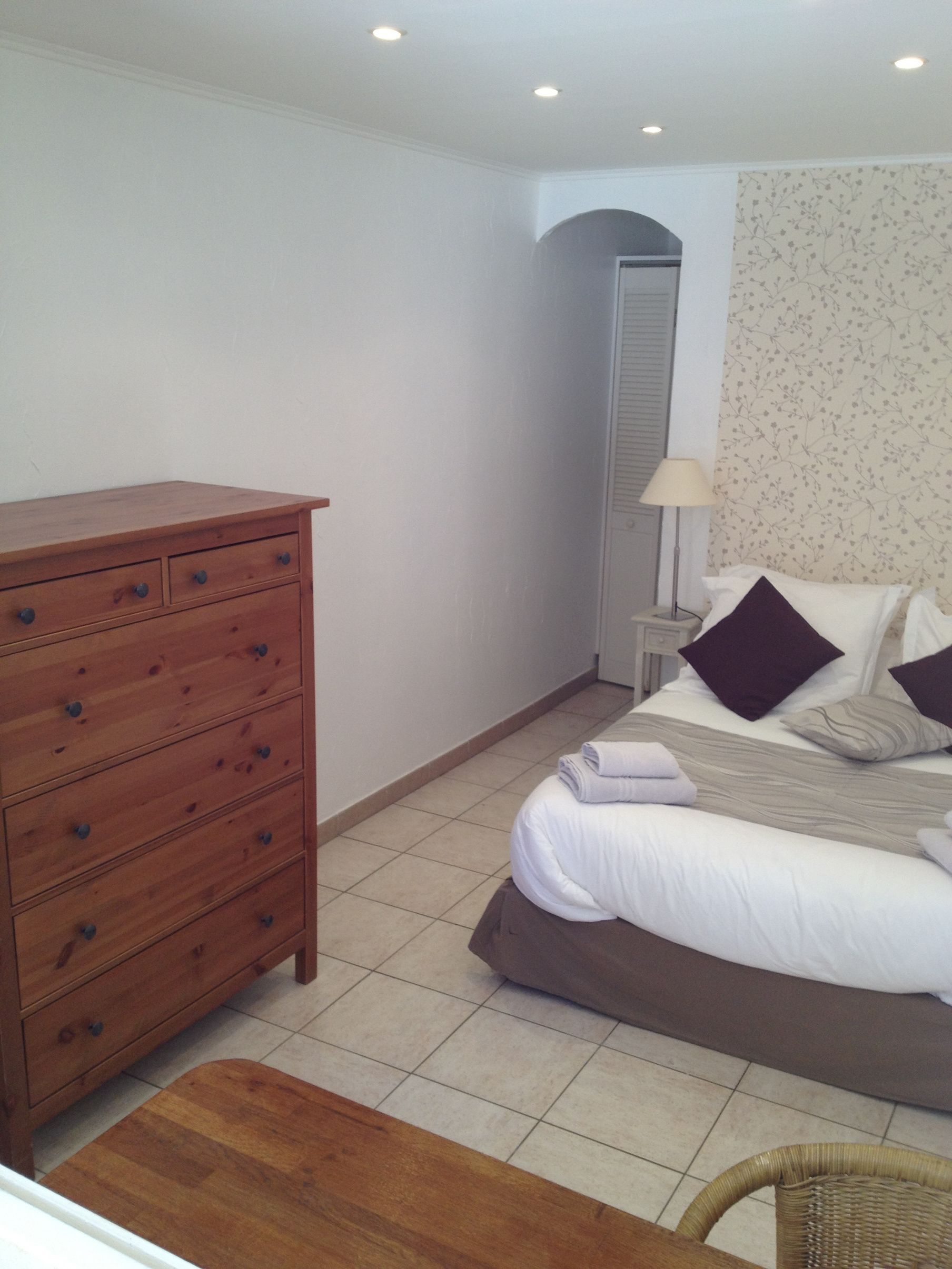 Une chambre avec une commode en bois, un lit avec des oreillers, une table de chevet avec une lampe et une chaise.