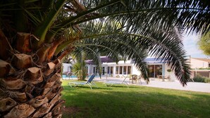 A pool area with lounge chairs, palm trees, and a building with a blue roof in the background. - Citotel Le Mirage (Istres)