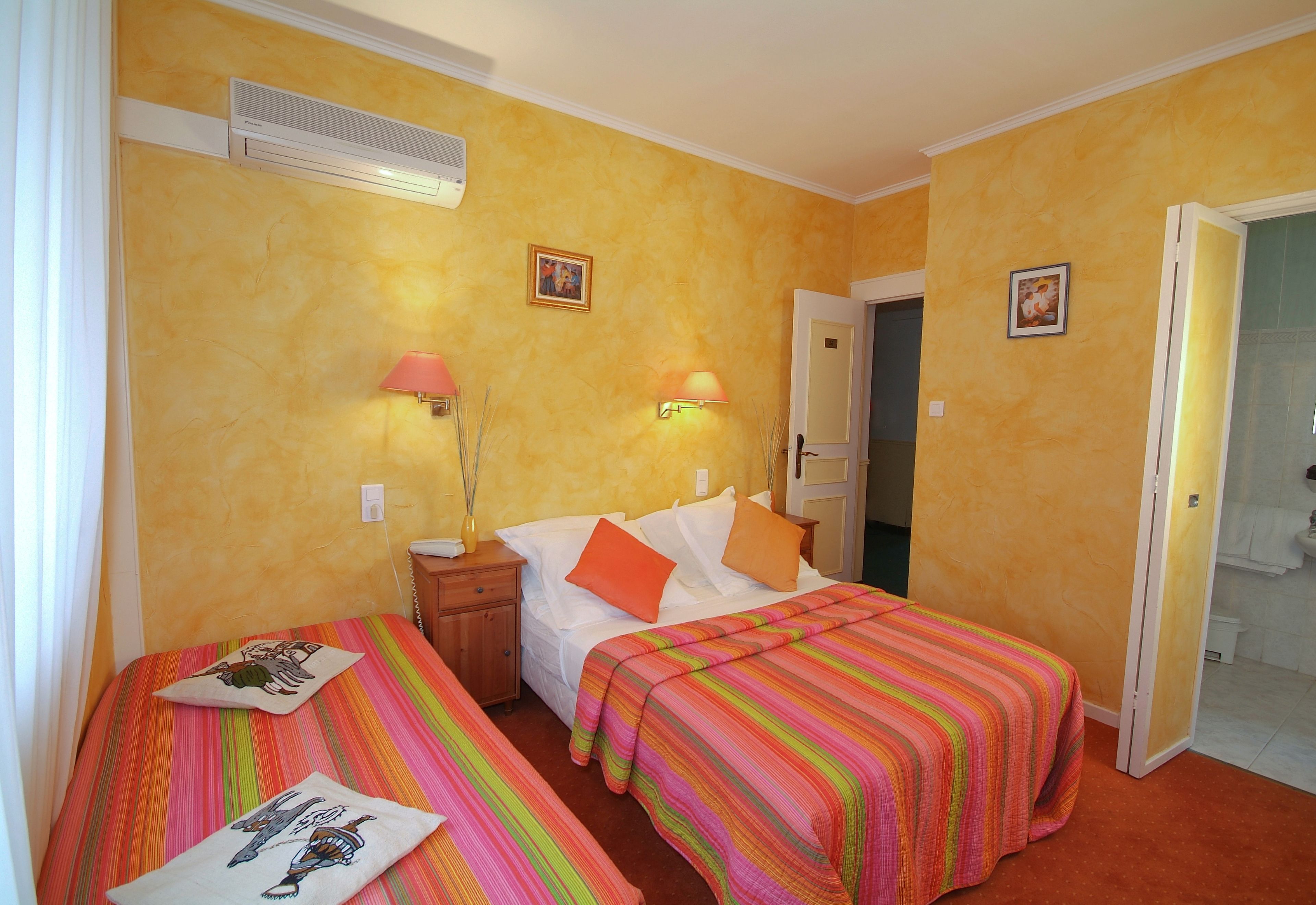A hotel room with two beds, each with a colorful striped bedspread and matching pillows. There are wall-mounted lamps on either side of the beds, and a small bedside table with a lamp and a vase.