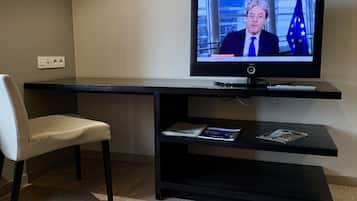 Una habitación moderna con una televisión de pantalla plana sobre un soporte negro, una silla blanca y un estante pequeño con revistas.