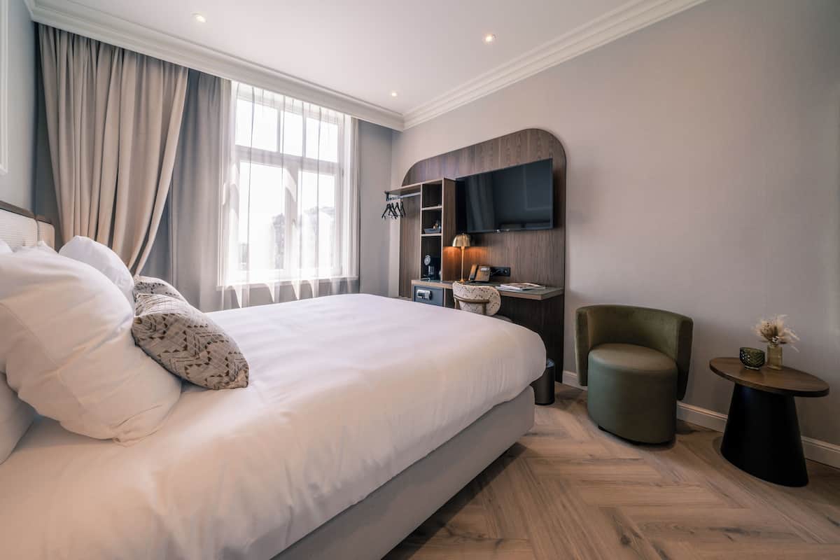 A modern hotel room with a large bed, a TV mounted on the wall, a desk with a lamp, and a small round table.
