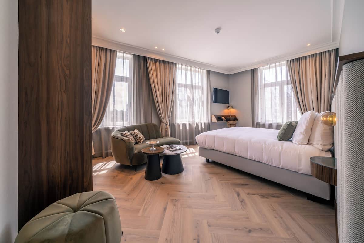 A modern hotel room with a large bed, a sofa, two side tables, and a TV mounted on the wall.