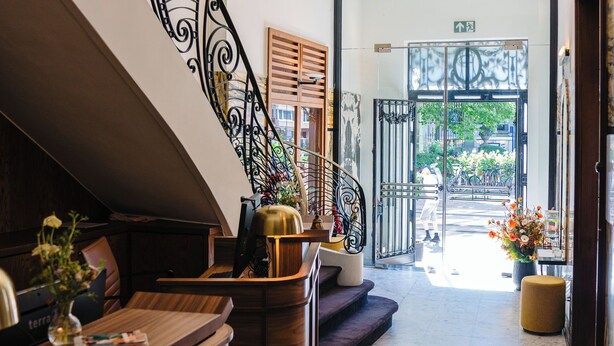 Un hall d’hôtel doté d’un comptoir d’accueil en bois, d’un escalier en colimaçon orné de rampes en métal ouvragé, et offrant une vue sur la rue.
