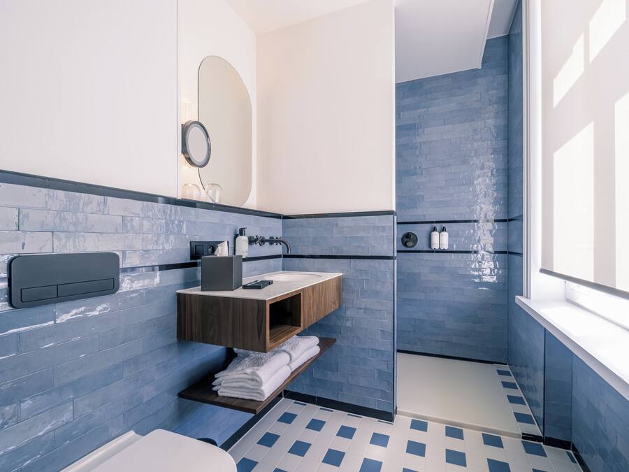 Une salle de bain moderne avec une douche aux carreaux bleus, un meuble-lavabo en bois, un miroir et une fenêtre laissant entrer la lumière naturelle.