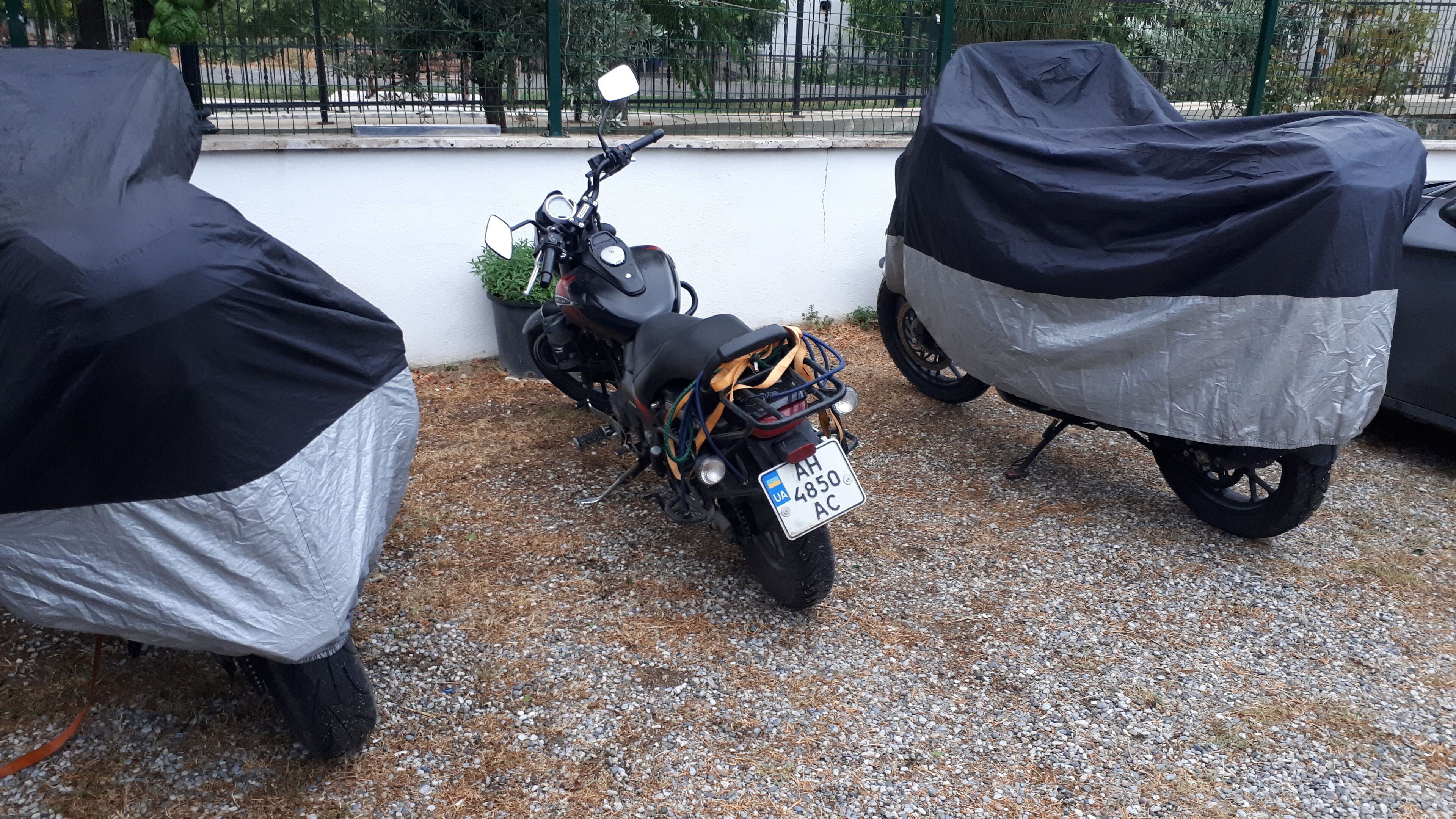 Two motorcycles covered with protective covers parked on a gravel surface.