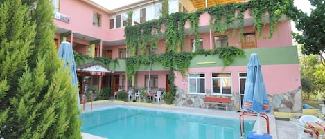 A pool area with a building featuring a pink facade and stone accents, surrounded by greenery and outdoor seating.