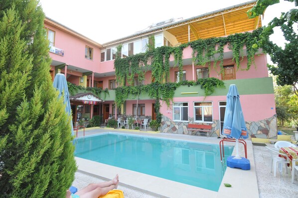A pool area with a building featuring a pink facade and stone accents, surrounded by greenery and outdoor seating. - Hotel Pamukkale (Denizli)