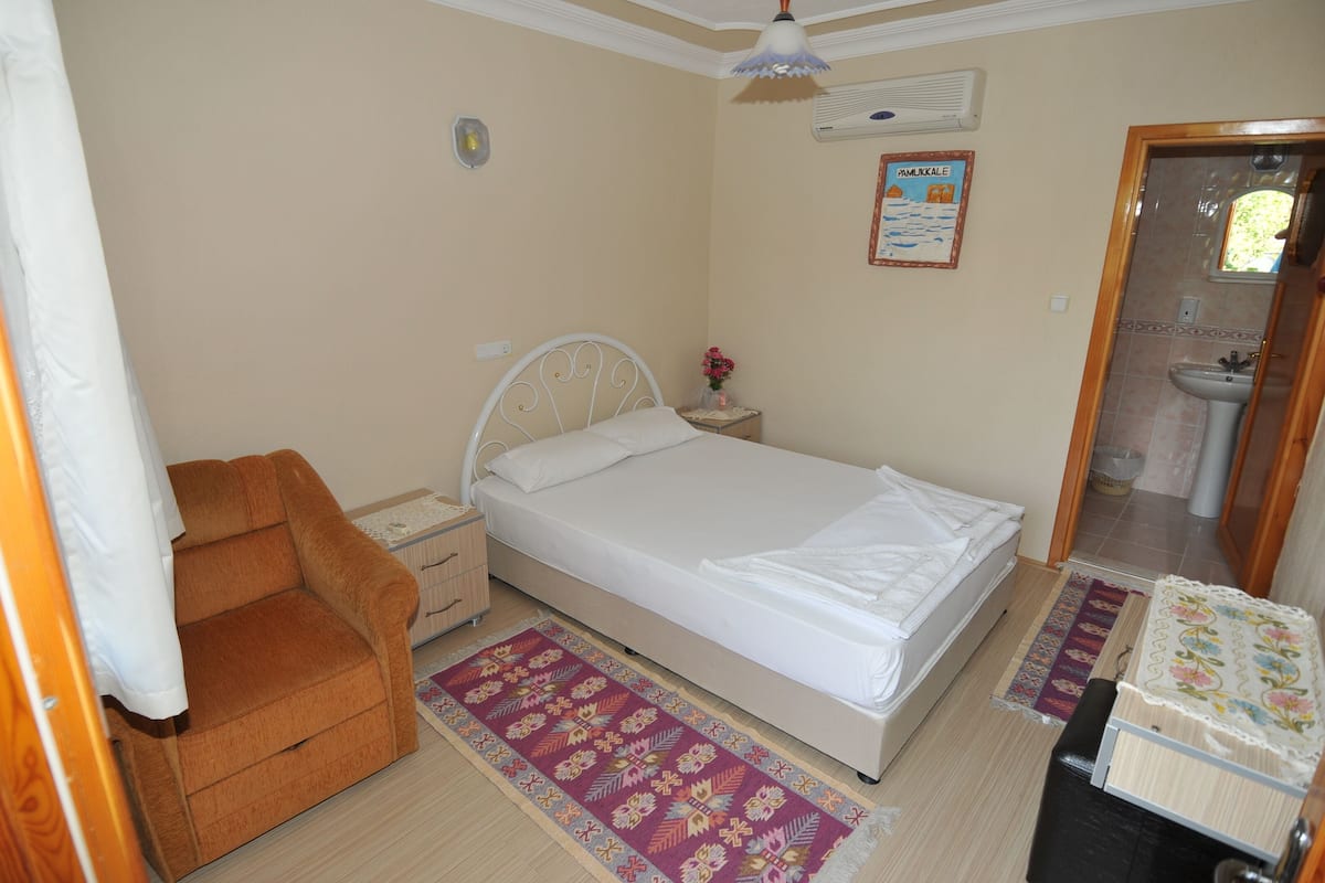A bedroom with a single bed, a nightstand, a chair, a rug, and a bathroom visible through an open door.