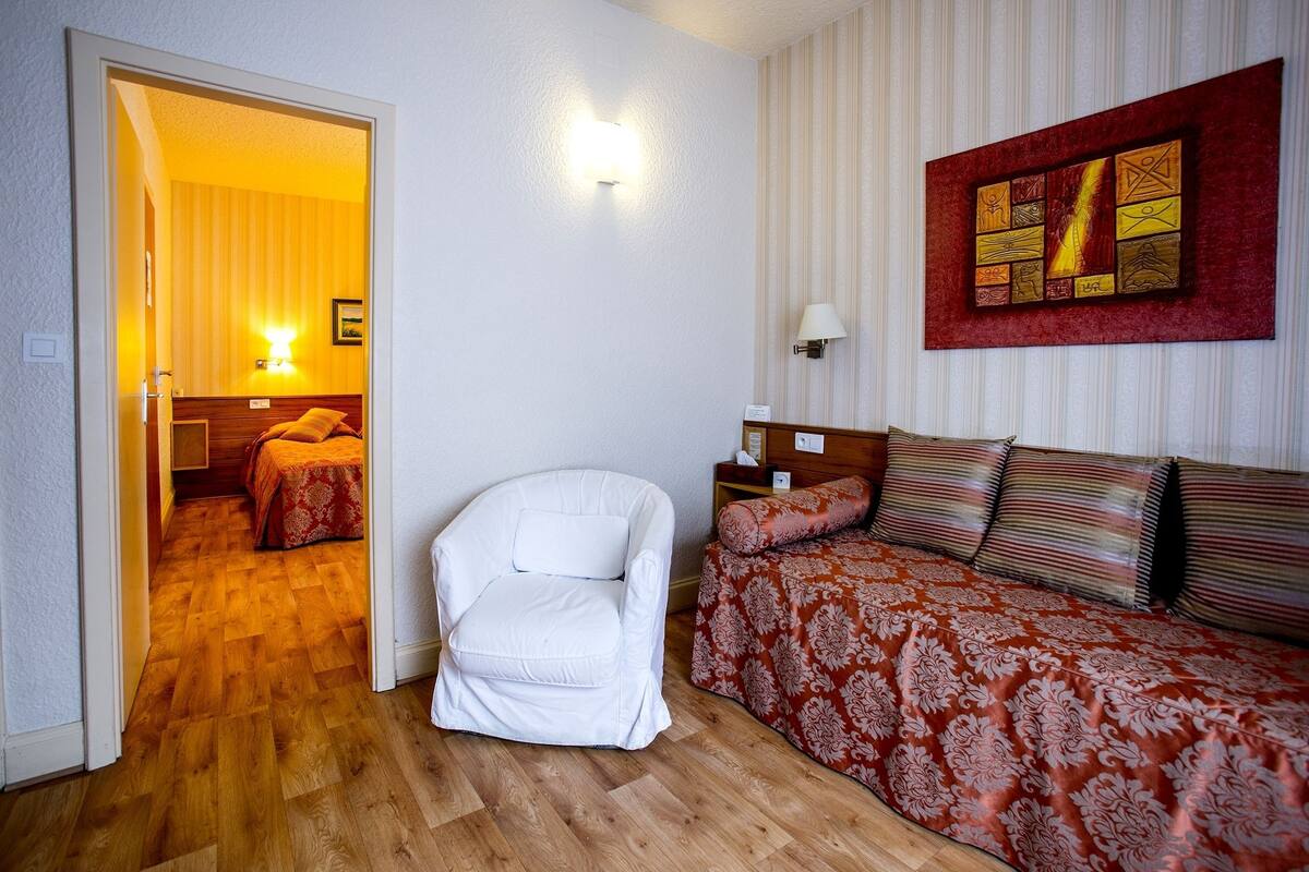 A hotel room with a wooden floor, a white armchair, a bed with red and white bedding, a nightstand with a lamp, and a painting on the wall.