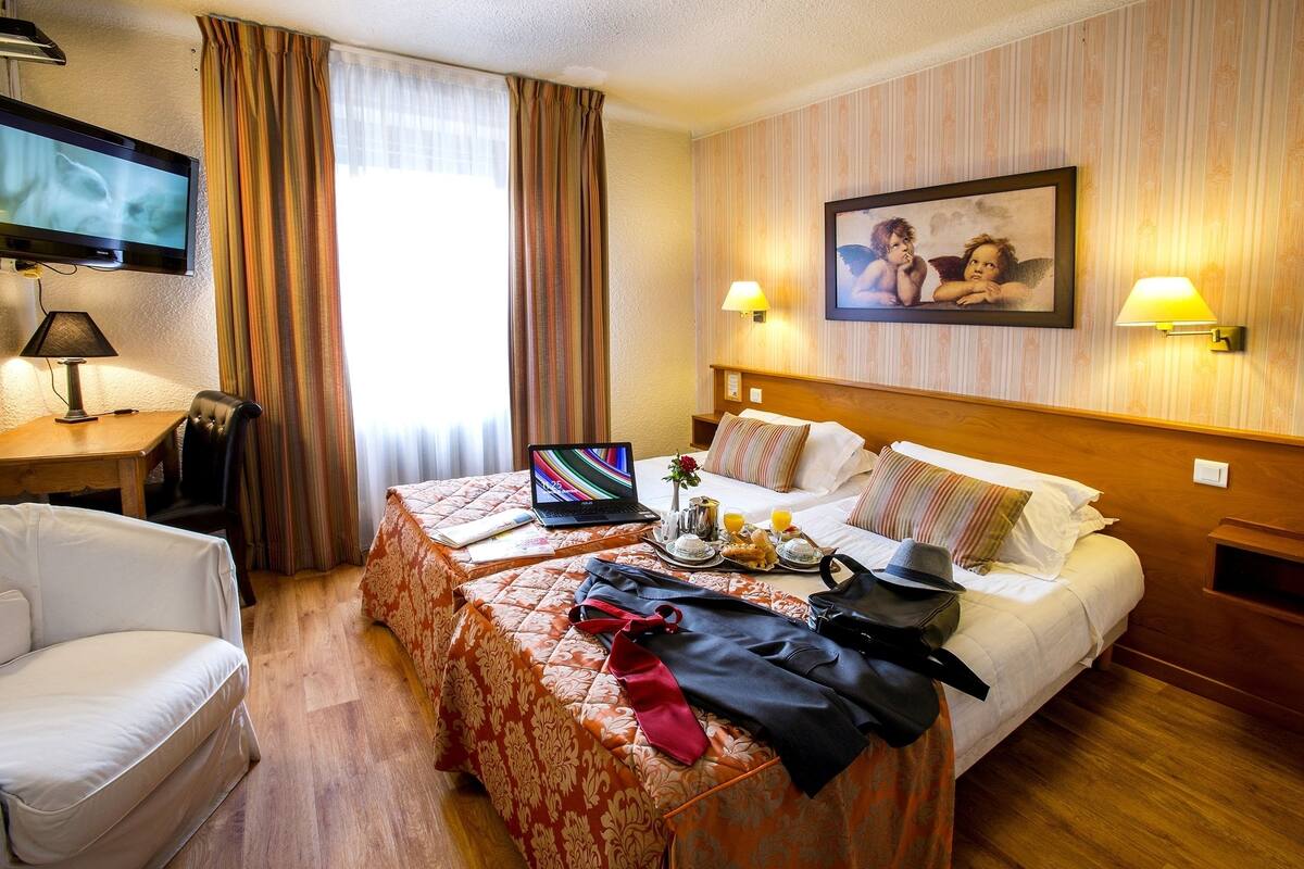 A hotel room with a bed, a desk with a laptop, a TV, and a painting on the wall.