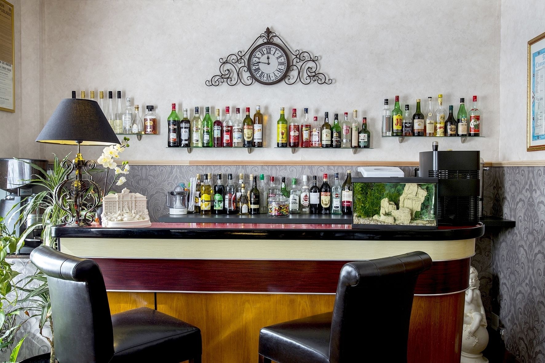 Uma área de bar com um relógio, diversas garrafas de bebidas alcoólicas, uma máquina de café e duas cadeiras.