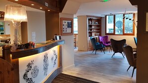 A reception area with a wooden counter, a lamp, and a framed bulletin board. There are chairs and a table in the background, and a window with a view of trees. - Logis Hôtel le Relais du Mont Blanc (Magland)