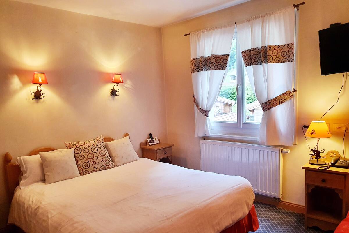 Un lit bien fait, agrémenté d’oreillers à motifs, une table de chevet avec une lampe, une fenêtre avec des rideaux et un radiateur.