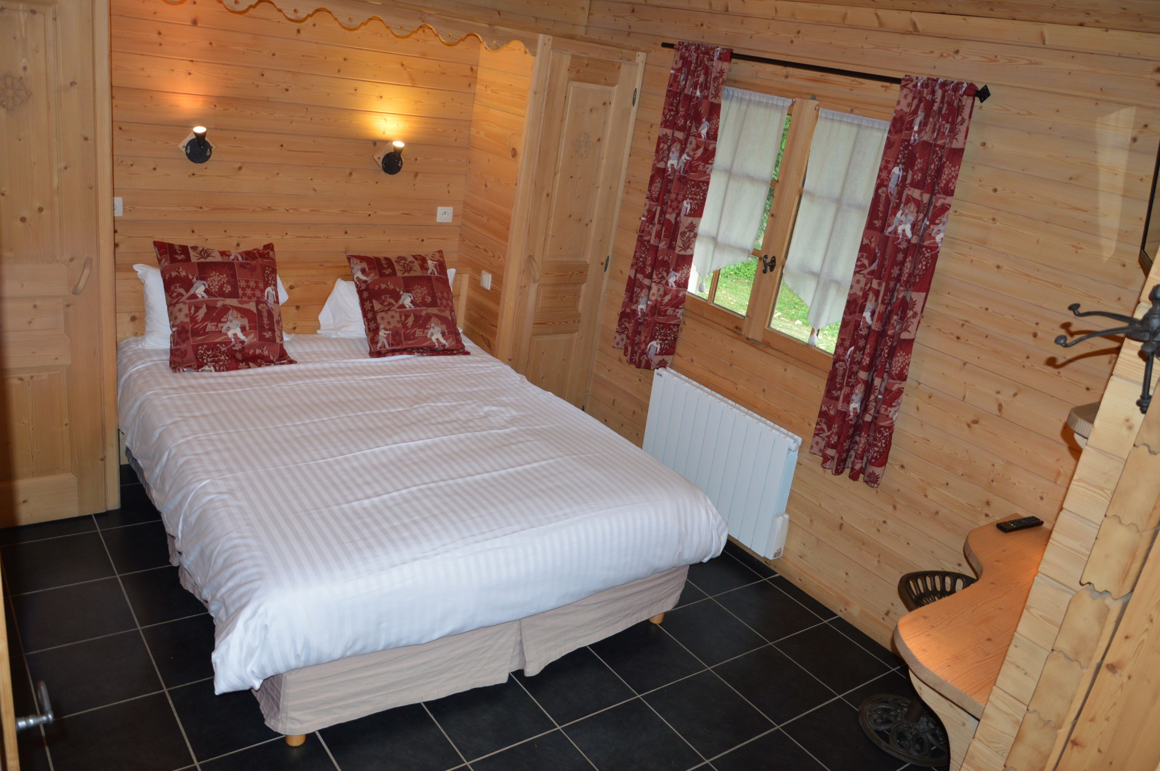 A wooden cabin bedroom with a bed, two pillows, a nightstand, a window with curtains, and a radiator.