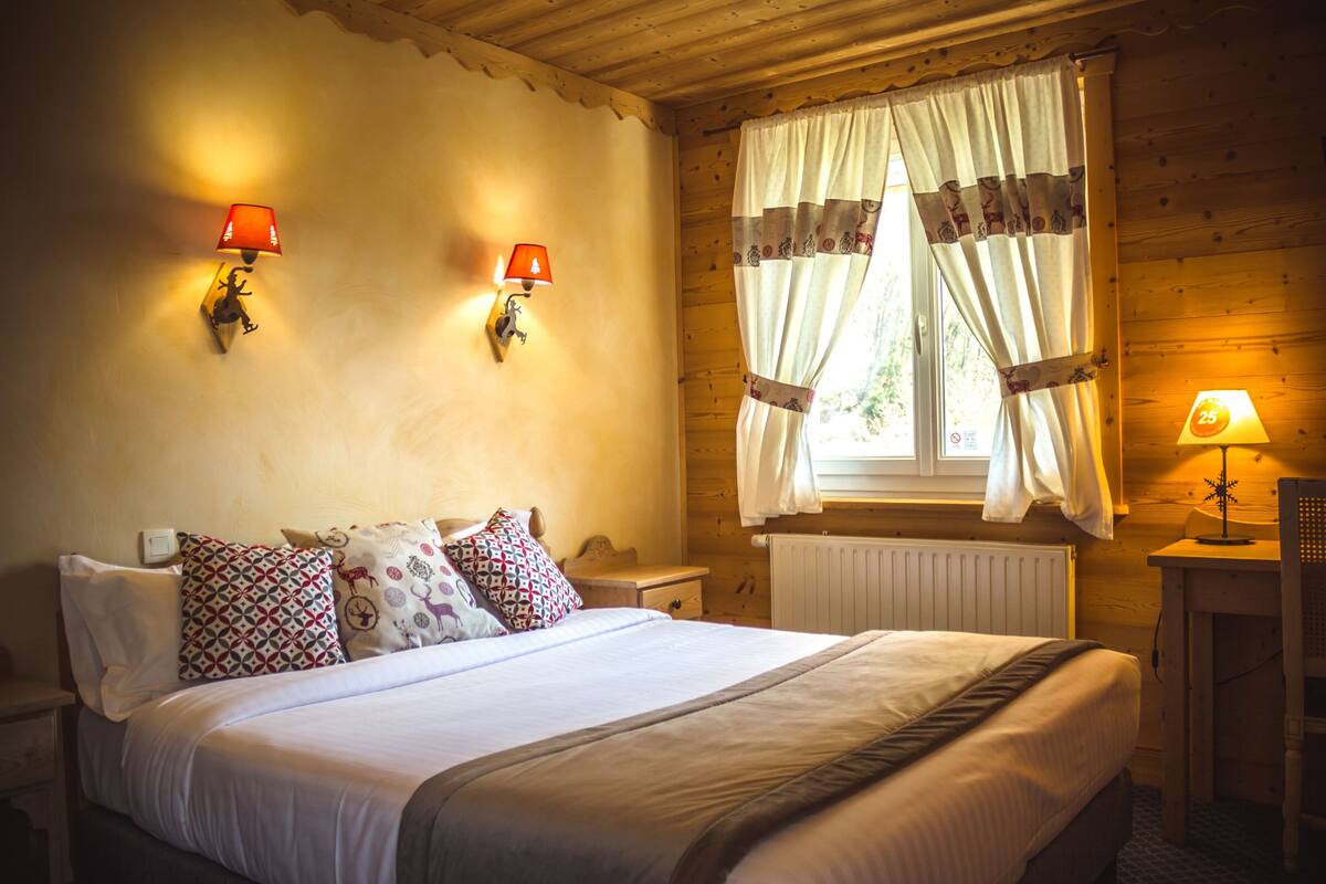 Une pièce chaleureuse en bois, avec un lit, deux lampes fixées au mur, une fenêtre avec des rideaux et une petite table sur laquelle se trouve une lampe.