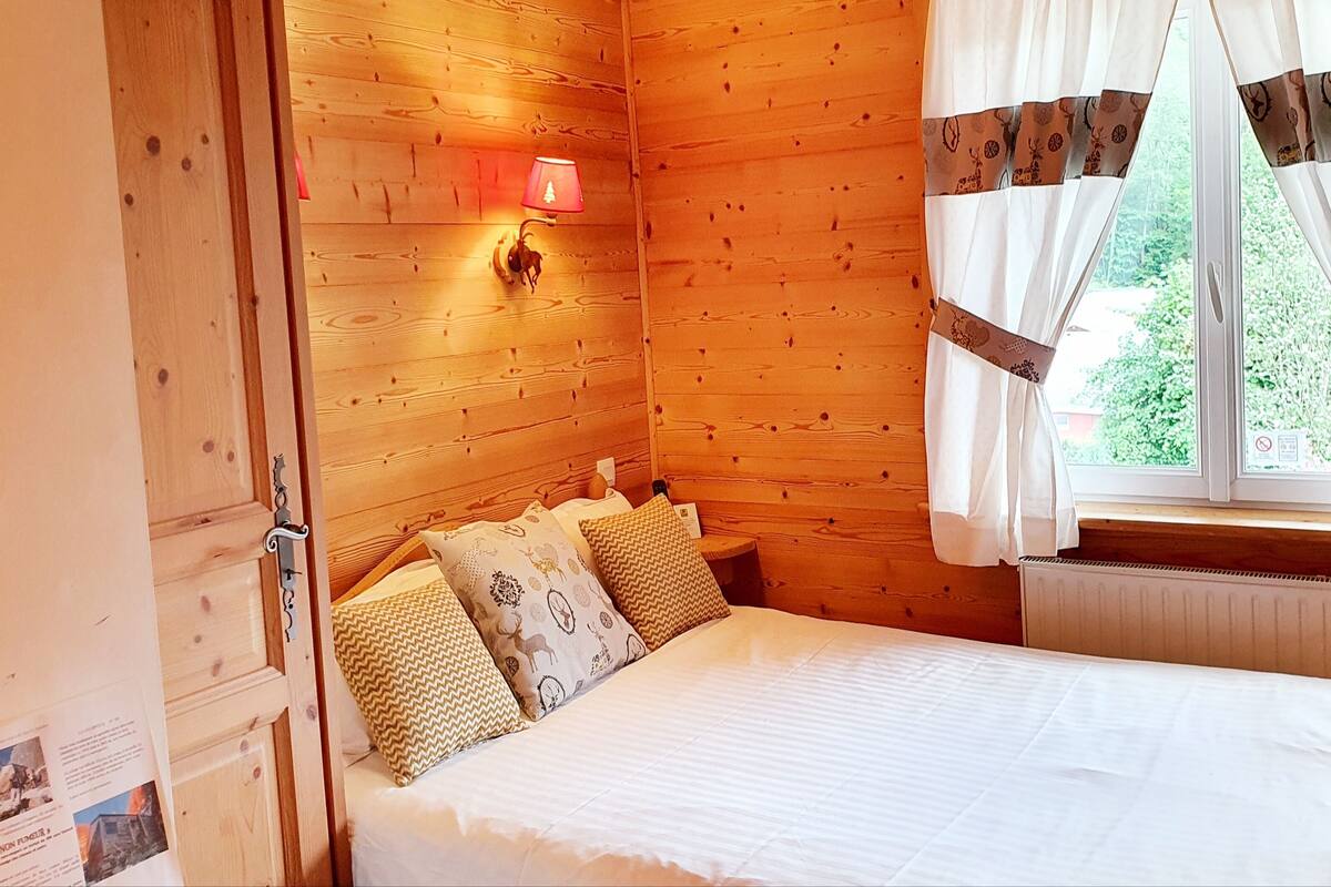 Une pièce en bois avec un lit, une lampe fixée au mur et une fenêtre avec des rideaux.