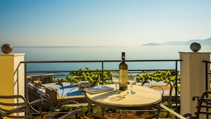 A terrace with a table, chairs, a bottle of wine, and a book, overlooking a calm sea and greenery.