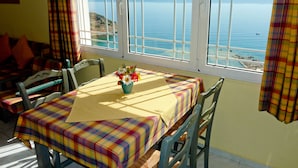 A dining area with a table set for two, a window with a view of the sea, and a chair with a plaid cover.