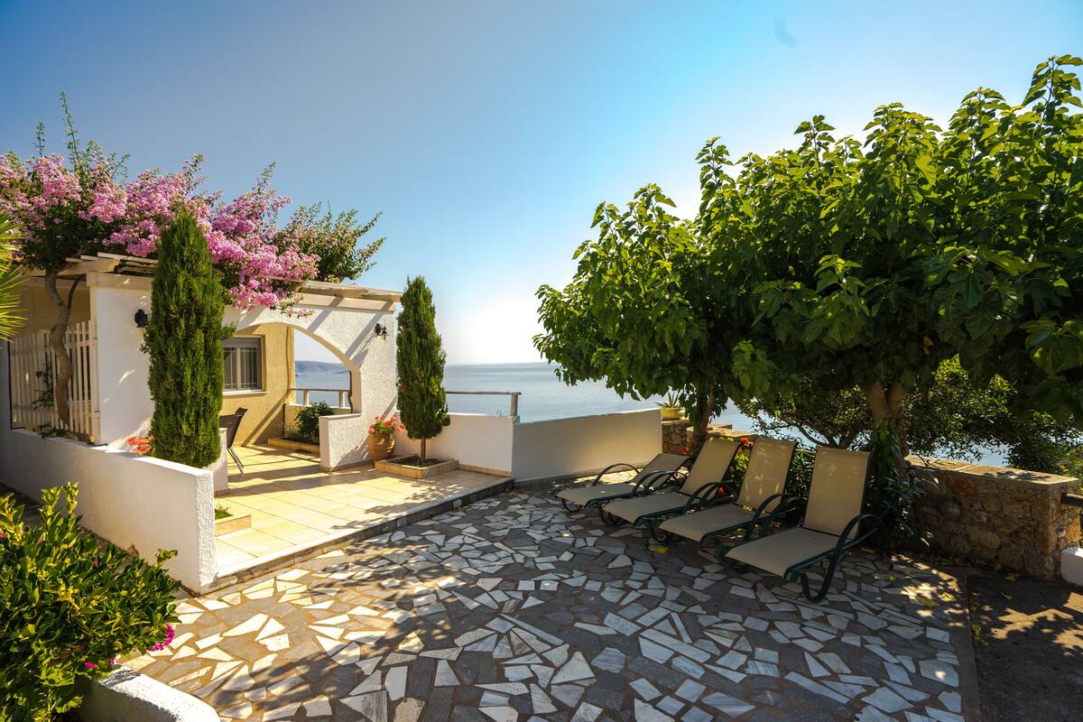 A sunlit patio with mosaic flooring, outdoor seating, and a view of the sea.