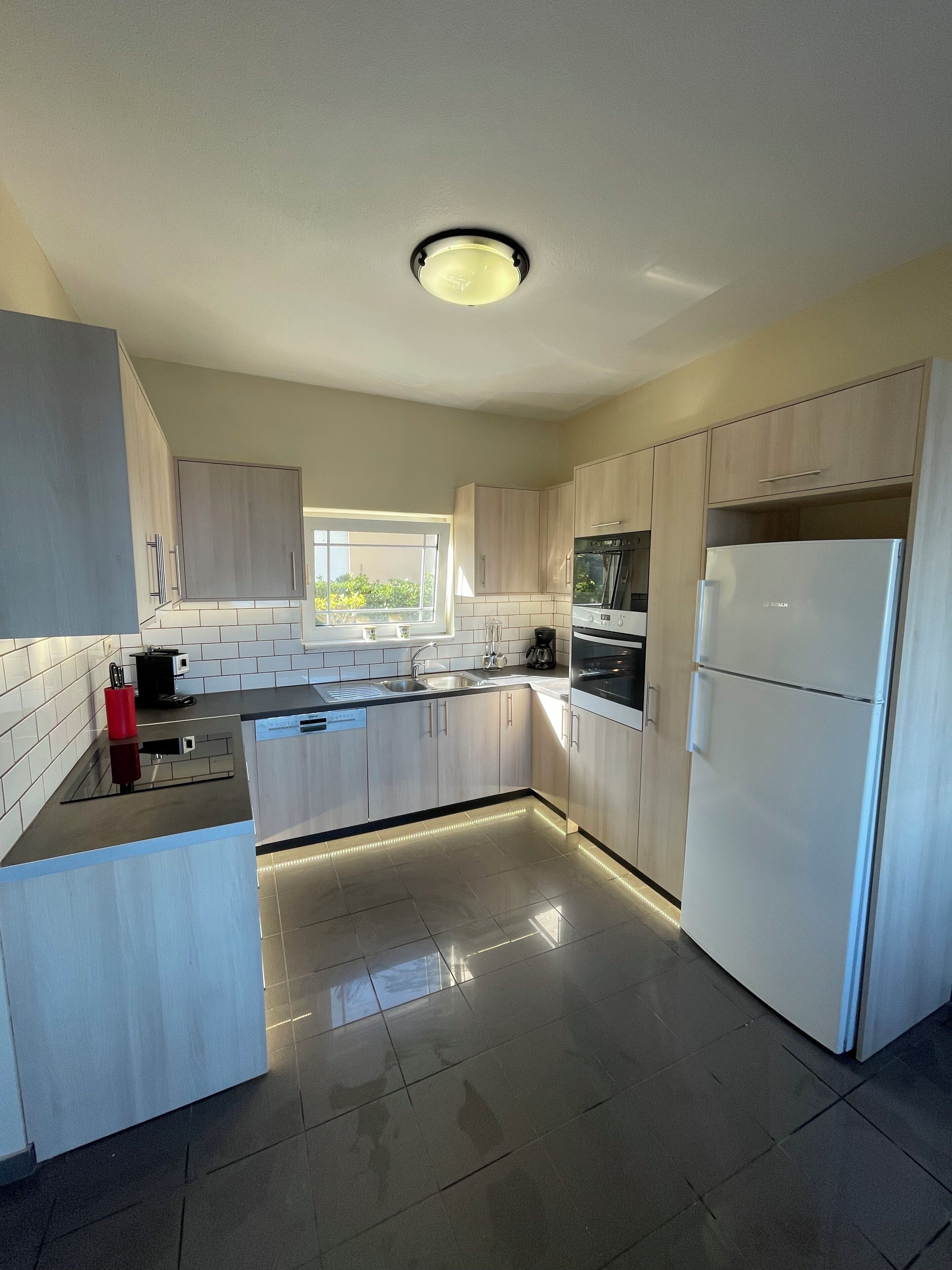A modern kitchen with light wood cabinets, stainless steel appliances, and a large window providing natural light.