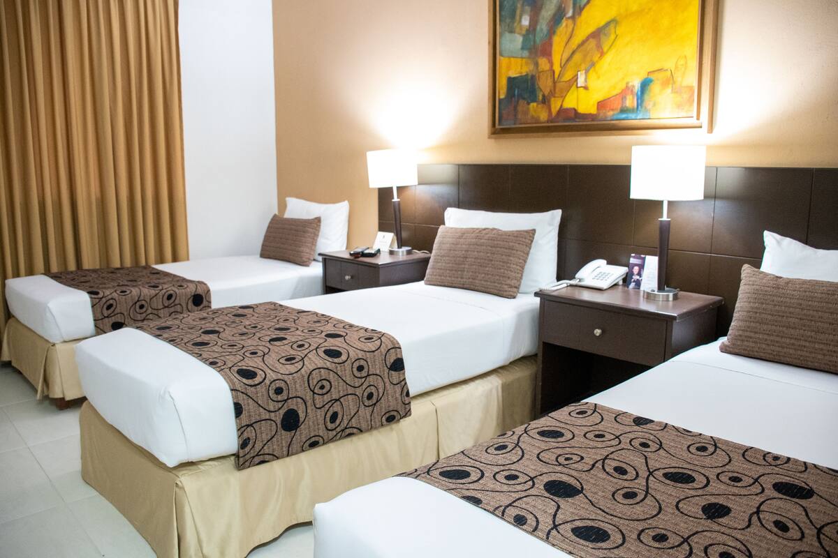 Ein Hotelzimmer mit drei Betten, jedes mit einer gemusterten Tagesdecke, weißer Bettwäsche und einem Nachttisch mit Lampe.
