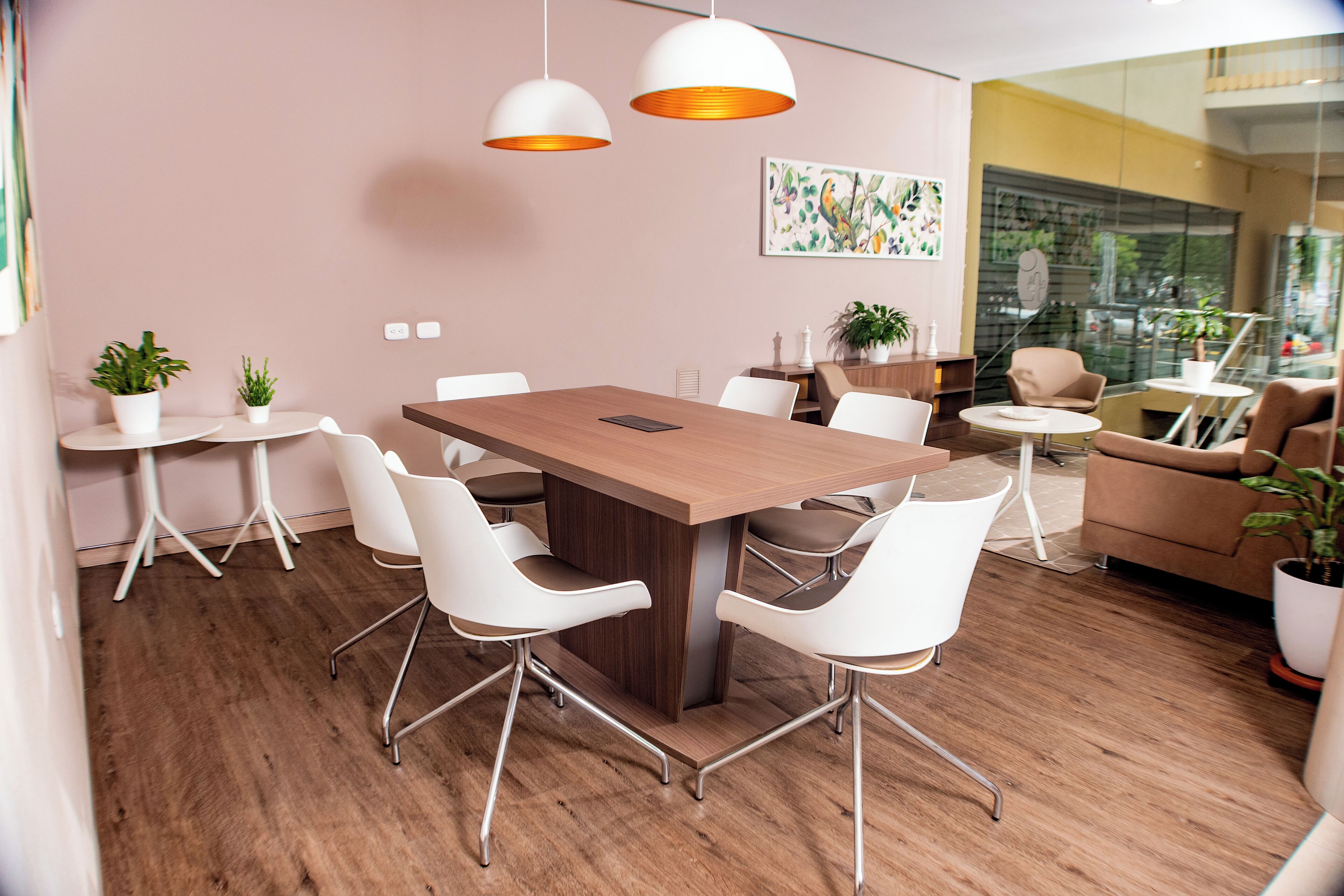 Una sala de reuniones moderna con una mesa de madera, sillas blancas, lámparas colgantes y plantas en macetas.