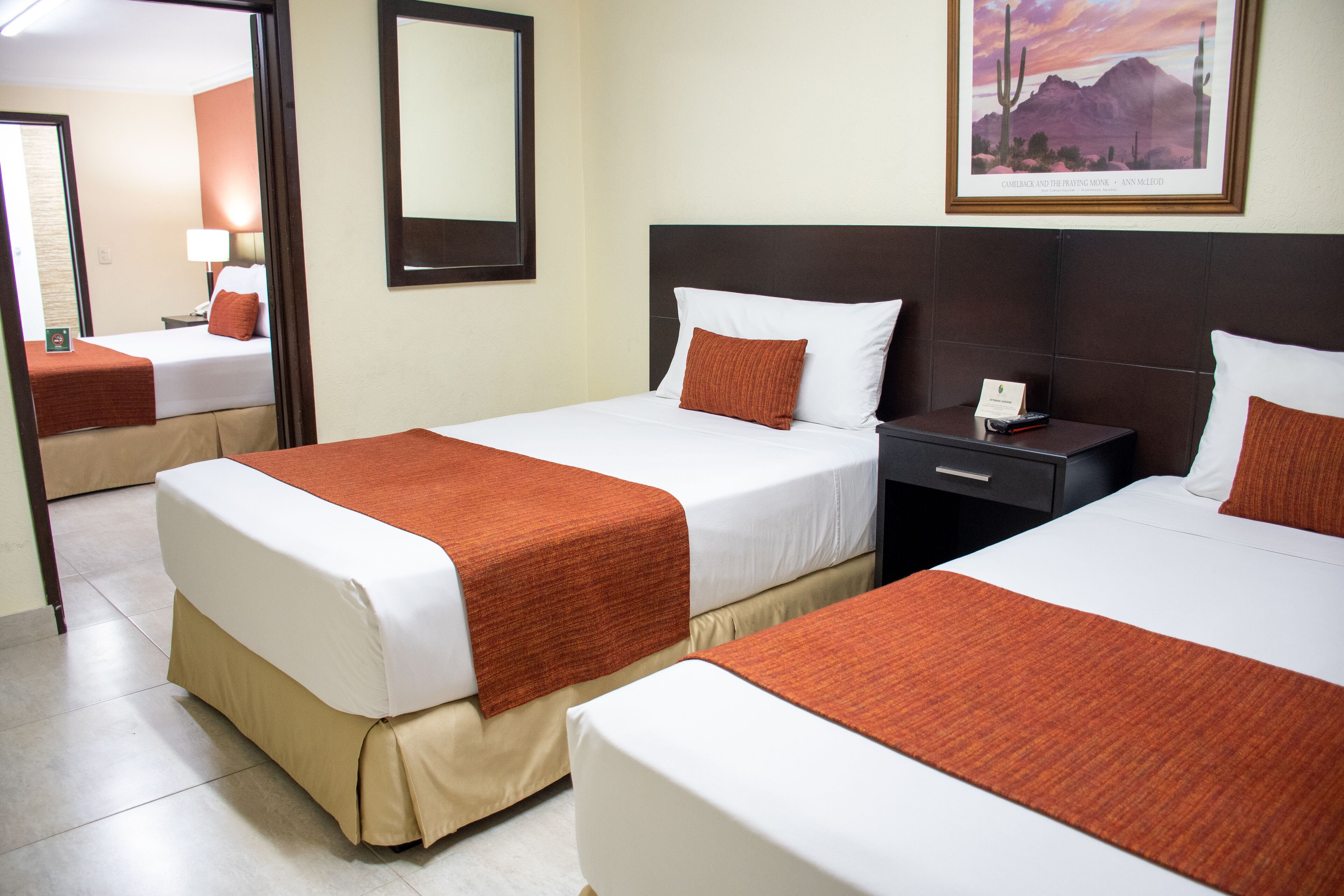 A hotel room with two beds, each with a white mattress and orange bedding. There is a nightstand with a phone and a framed picture on the wall.