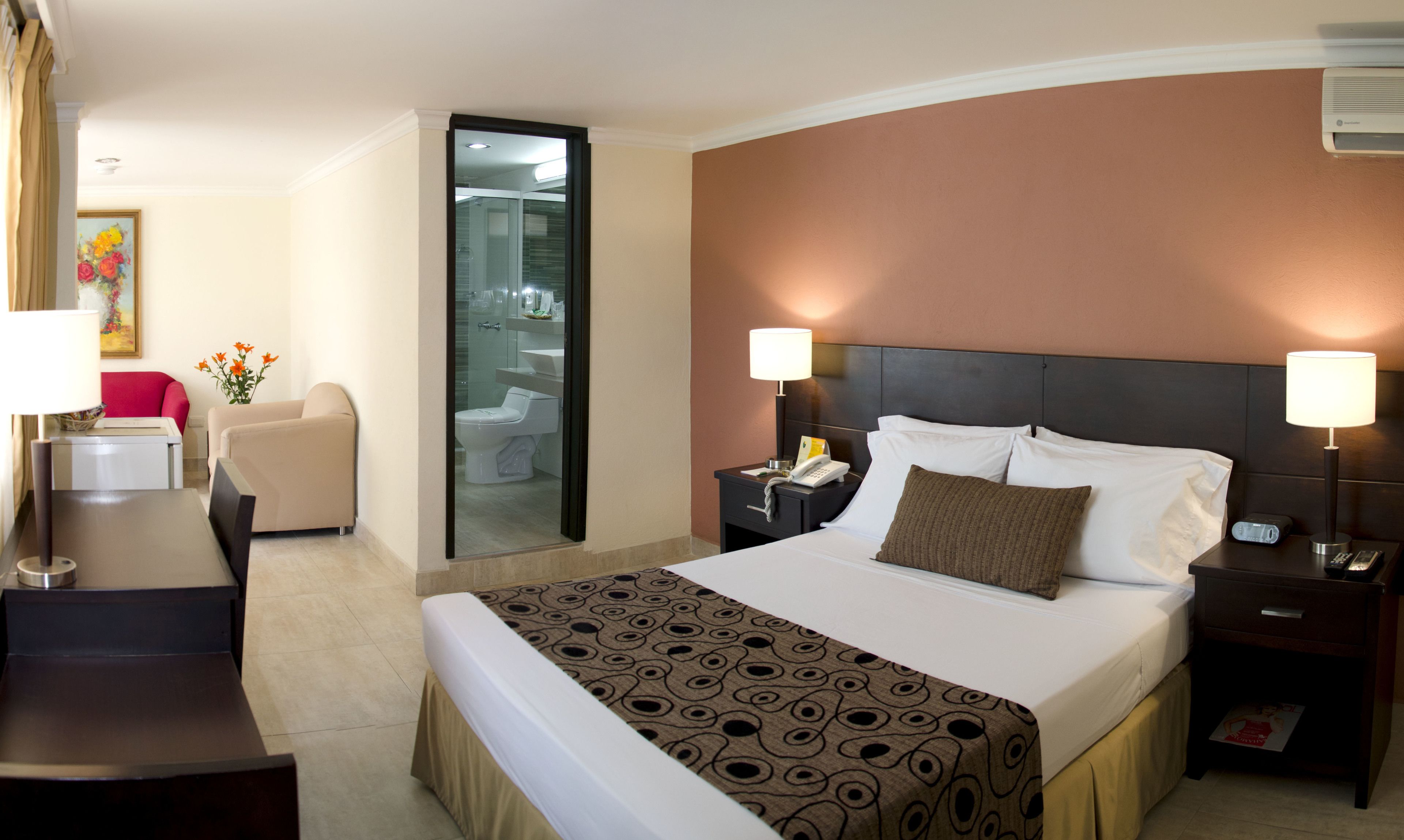 Ein Hotelzimmer mit Bett, Nachttischen, Schreibtisch, Stuhl und einem durch eine Glastür einsehbaren Badezimmer.