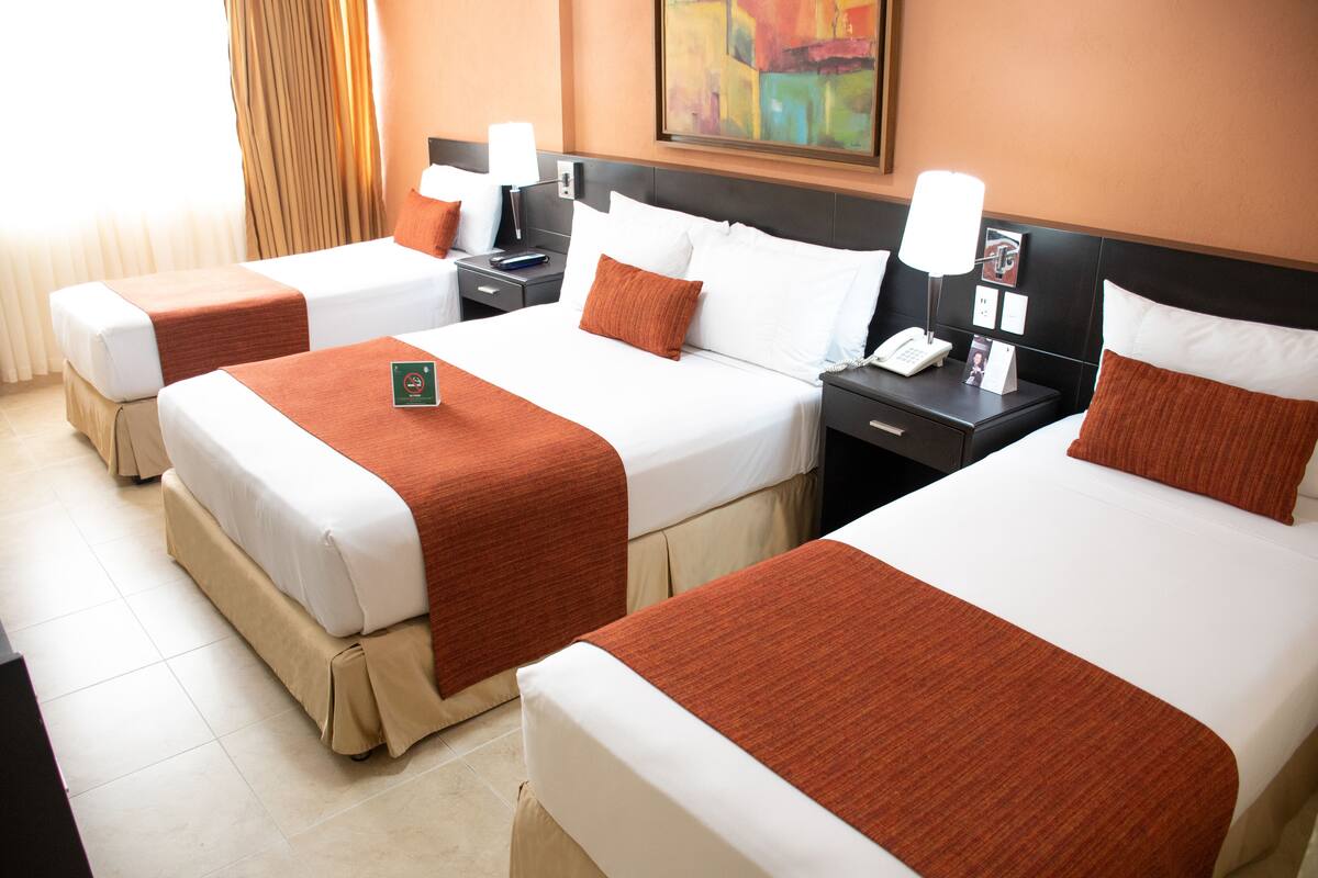 Ein Hotelzimmer mit drei Betten, jeweils mit weißen Laken und braunen Kopfteilen. An den Nachttischen stehen Lampen und ein Gemälde hängt an der Wand.