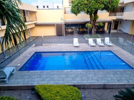 A rectangular swimming pool with a clear blue water surface, surrounded by a paved deck. There are white lounge chairs arranged around the pool, and a building with a yellow facade and balconies in the background.