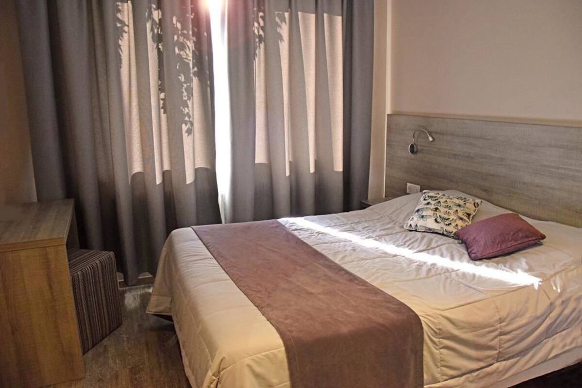 A hotel room with a bed, a wooden desk, and a window with curtains.