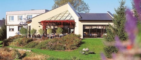 A building with a covered outdoor seating area, a pond with swans, and a grassy slope with plants.
