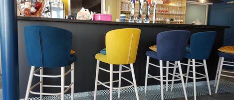Una zona de bar con cuatro taburetes, una barra con botellas y vasos, y un suelo estampado.