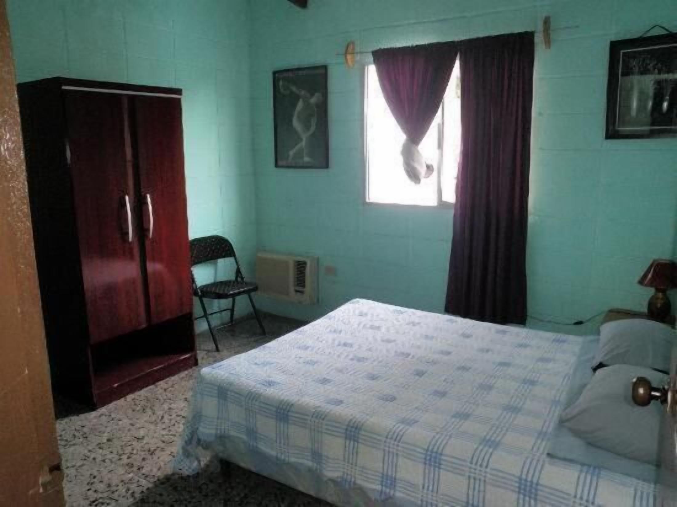 A bedroom with a bed, a wardrobe, a chair, a window with curtains, and a wall-mounted picture.