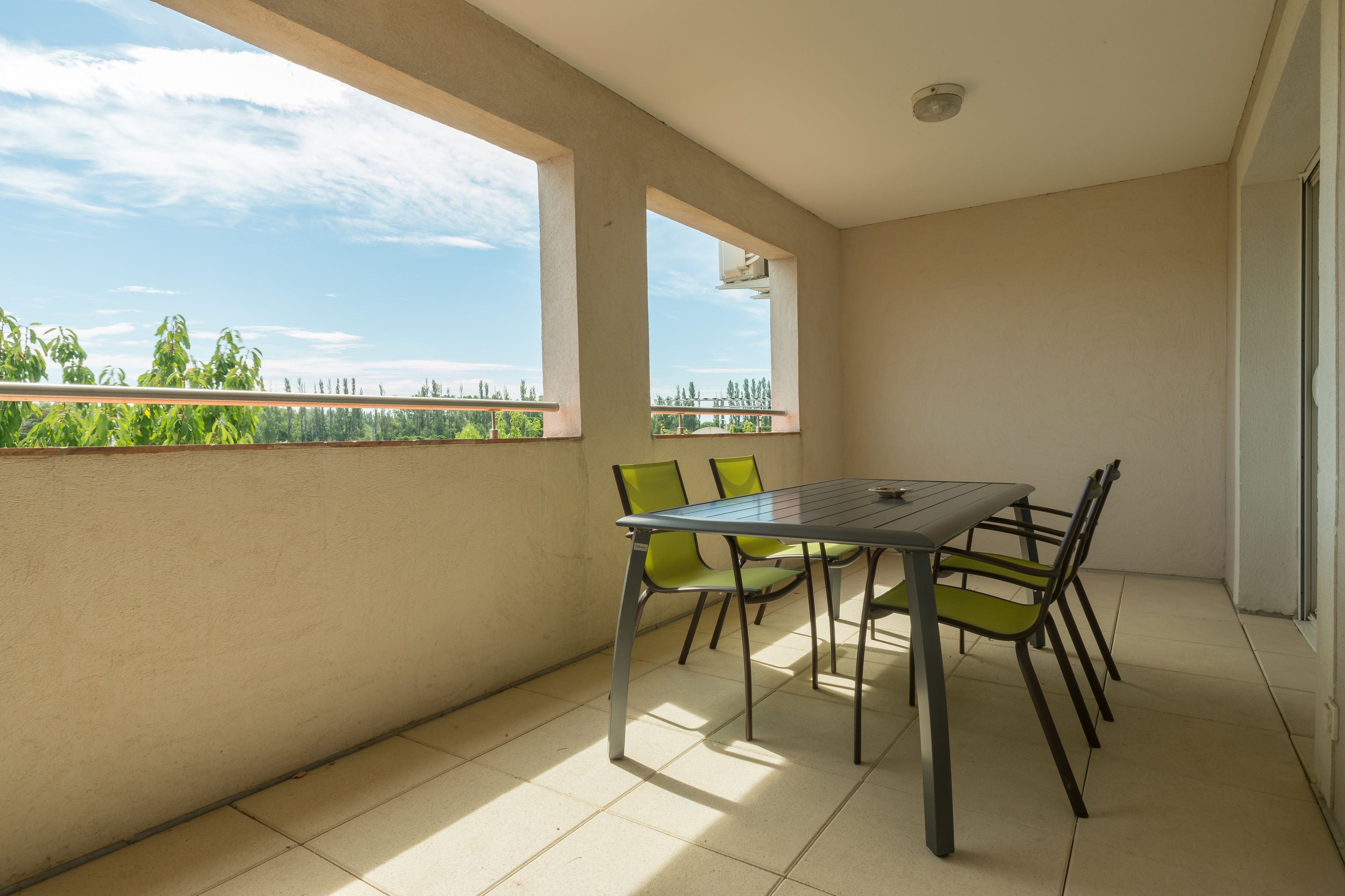 A balcony with a table and chairs, a view of greenery and trees, and a clear sky.