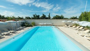A rectangular swimming pool with clear blue water, surrounded by white lounge chairs and umbrellas, and a concrete wall with greenery in the background.