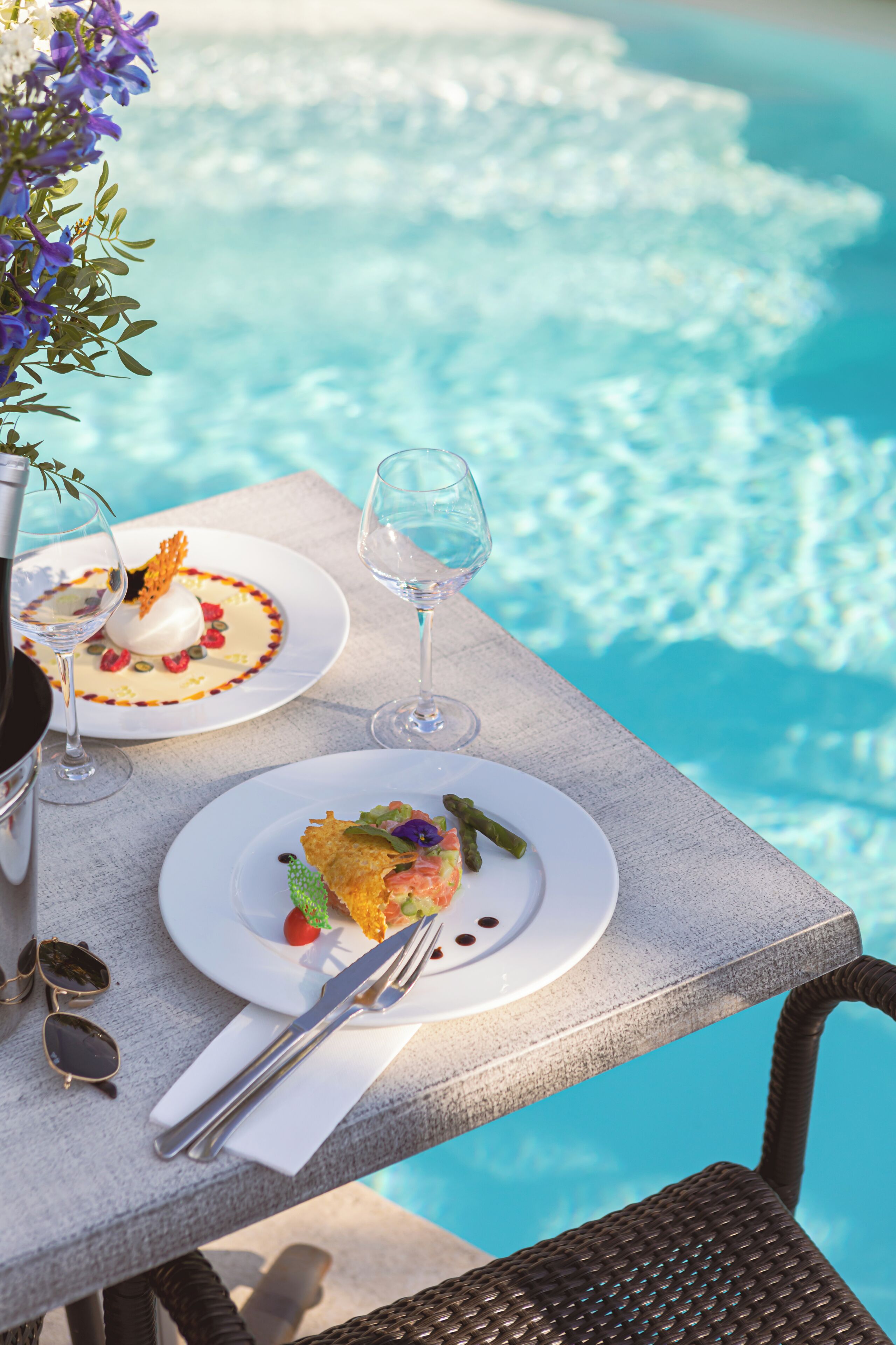 A dining table with plates of food, wine glasses, and cutlery, overlooking a pool.