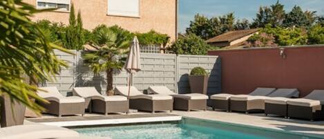 A swimming pool with lounge chairs and umbrellas, surrounded by a house and greenery.