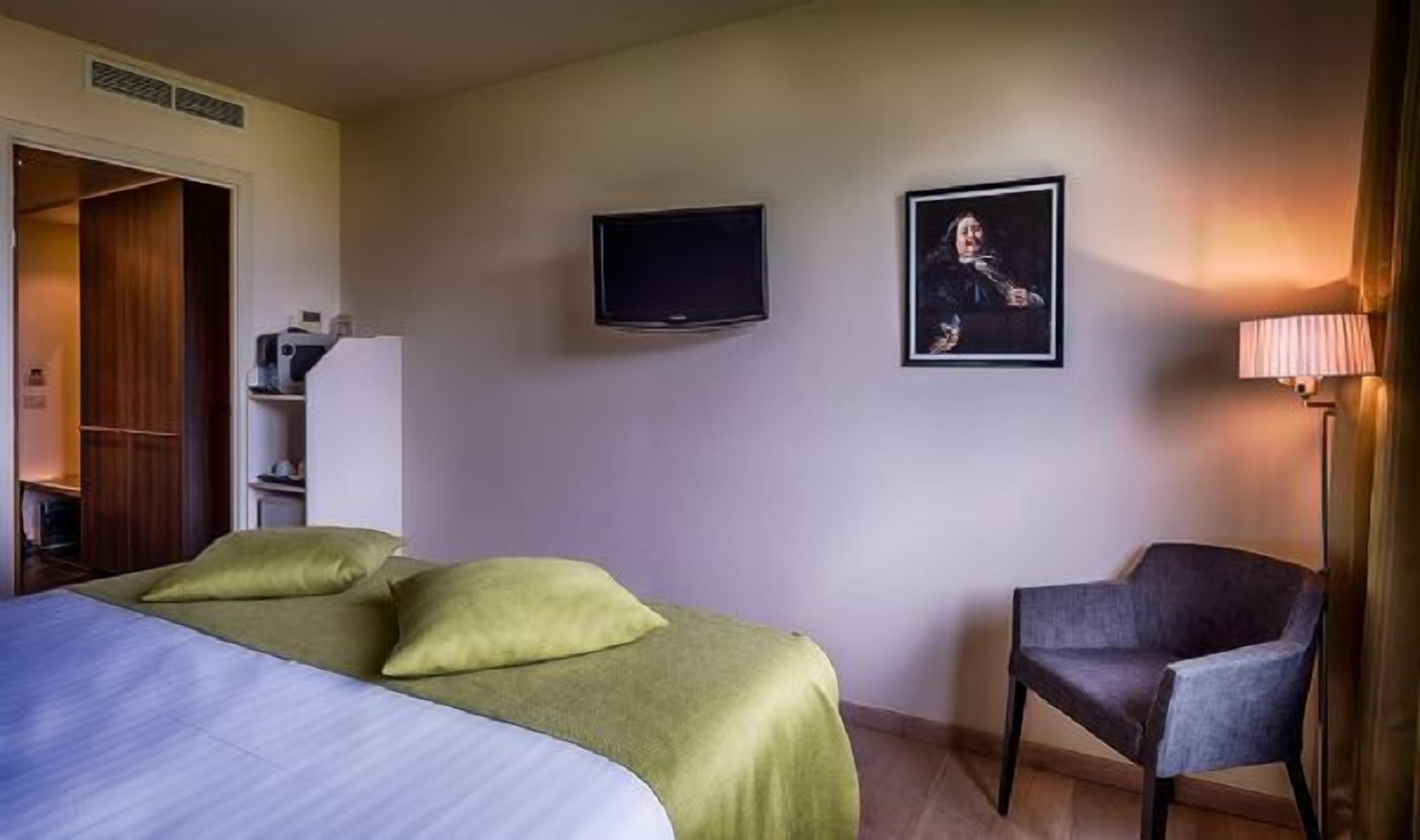 Hotellihuone, jossa on sänky, tuoli, televisio ja taulu seinällä.