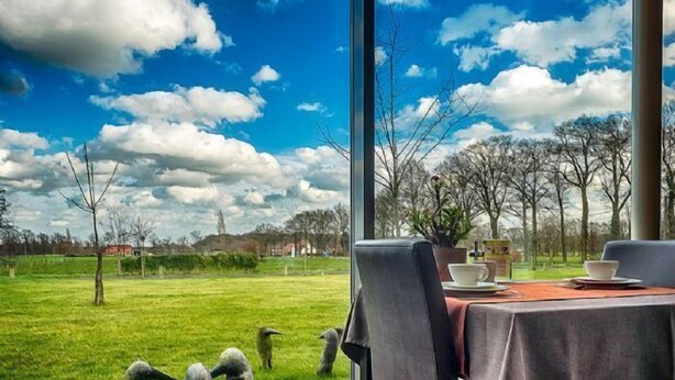 A dining area with a large window overlooking a grassy field and trees, with a clear blue sky and clouds. - Cleythil Hotel (Maldegem)