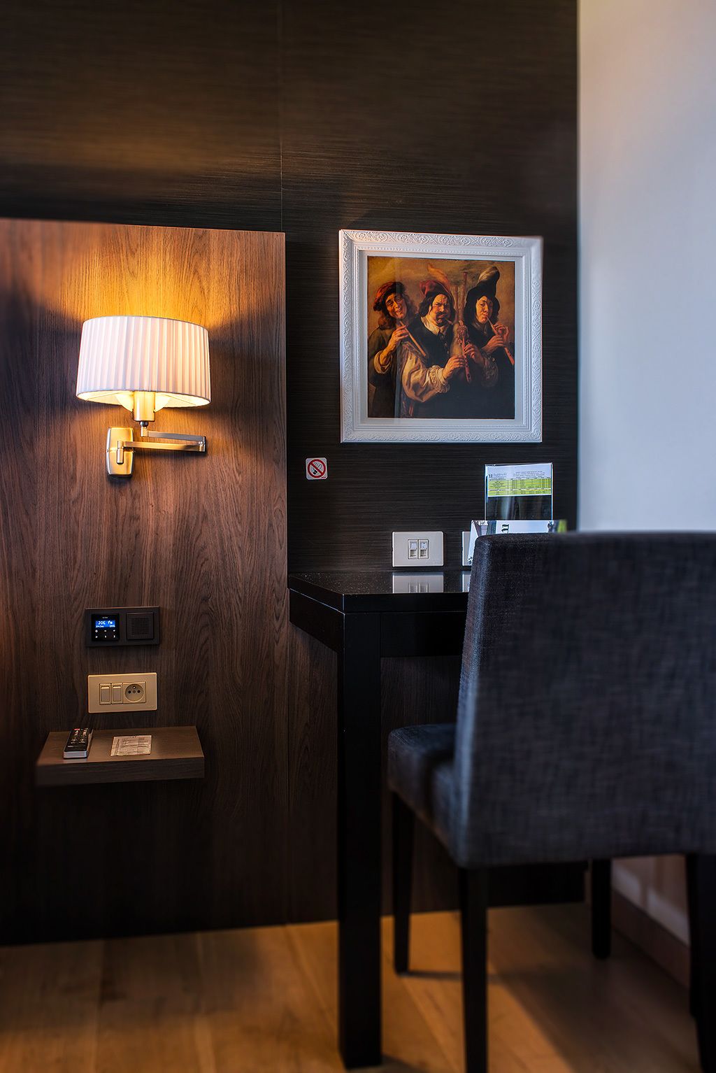 A hotel room with a wooden wall, a wall-mounted lamp, a framed painting, a desk with a chair, and a thermostat.