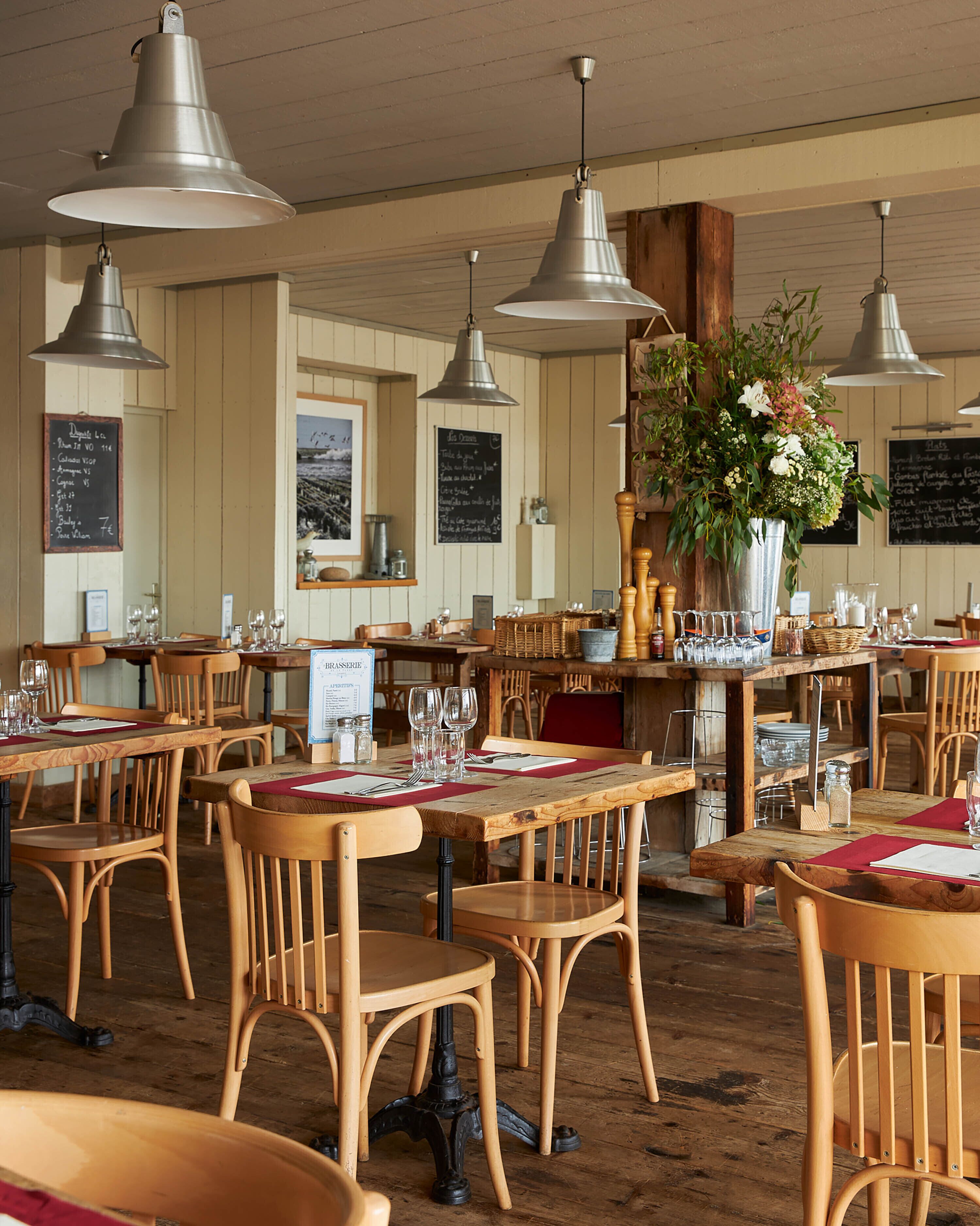 Een restaurantinterieur met houten tafels en stoelen, metalen hanglampen en een bloemstuk in het midden.