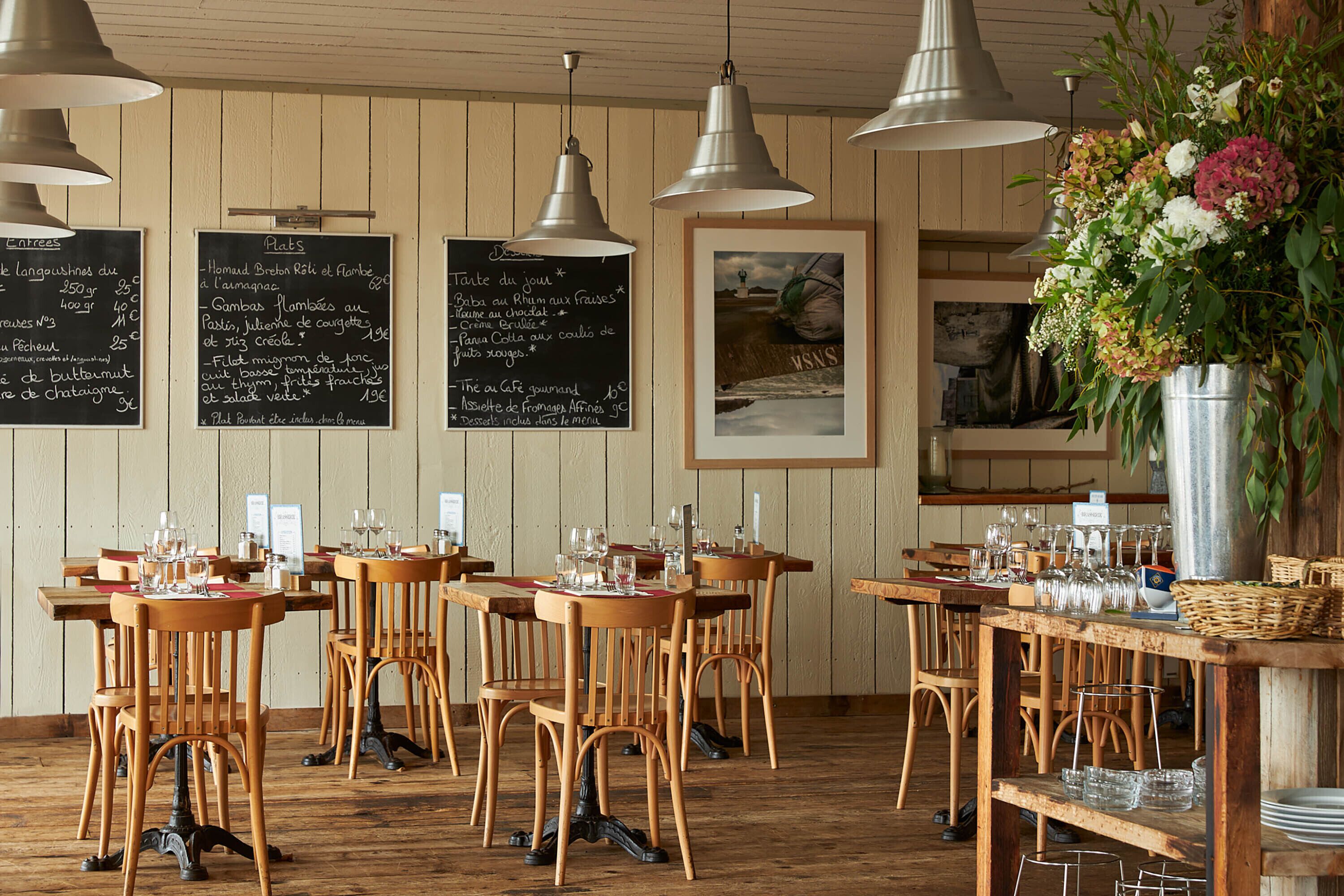 L’intérieur d’un restaurant avec des tables et des chaises en bois, des tableaux noirs affichant le menu, des suspensions décoratives et un arrangement floral.