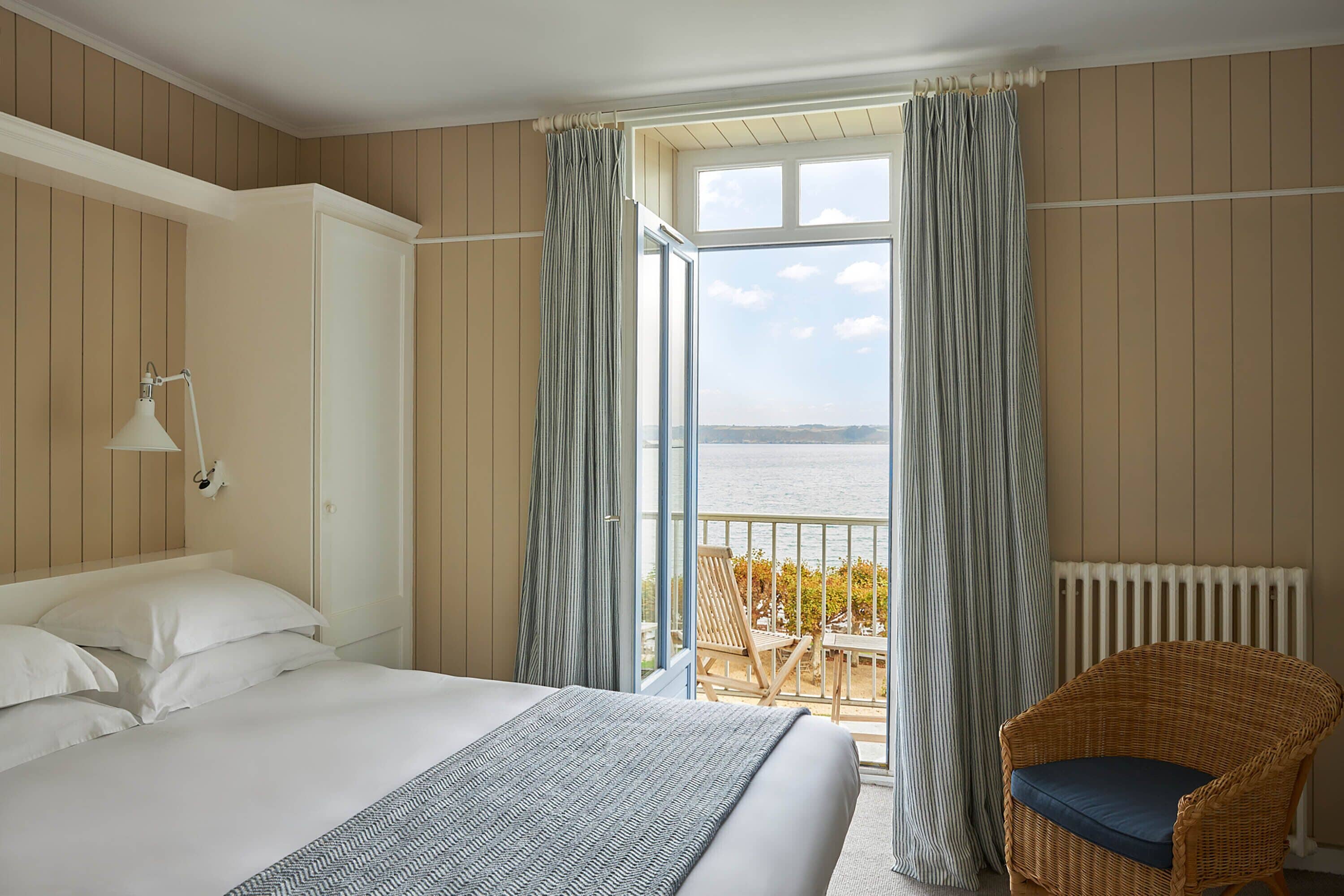 A hotel room with a large bed, a wicker chair, and a balcony with a view of the sea.